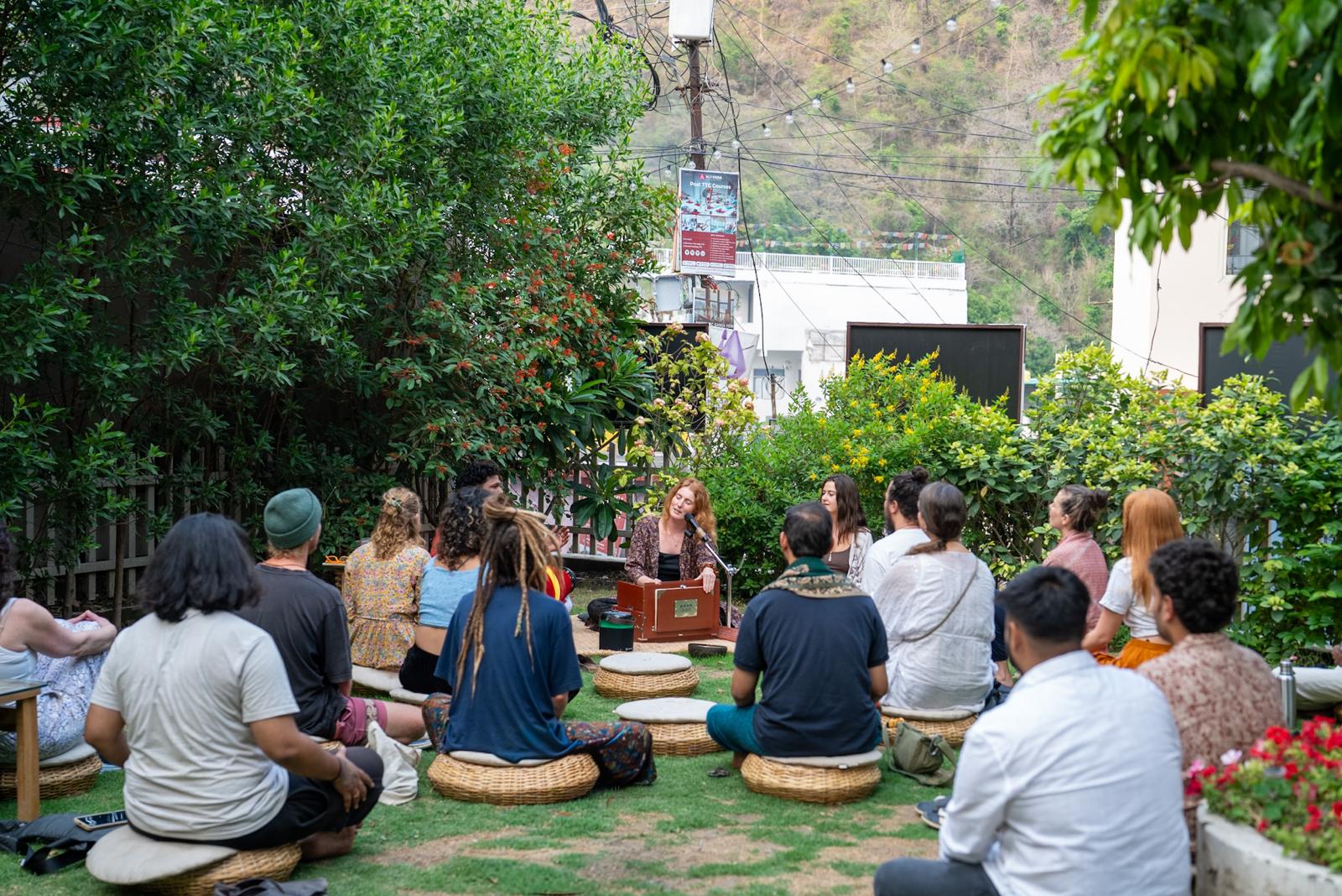A kirtan circle seated on cushions in a garden, harmonium at the centre