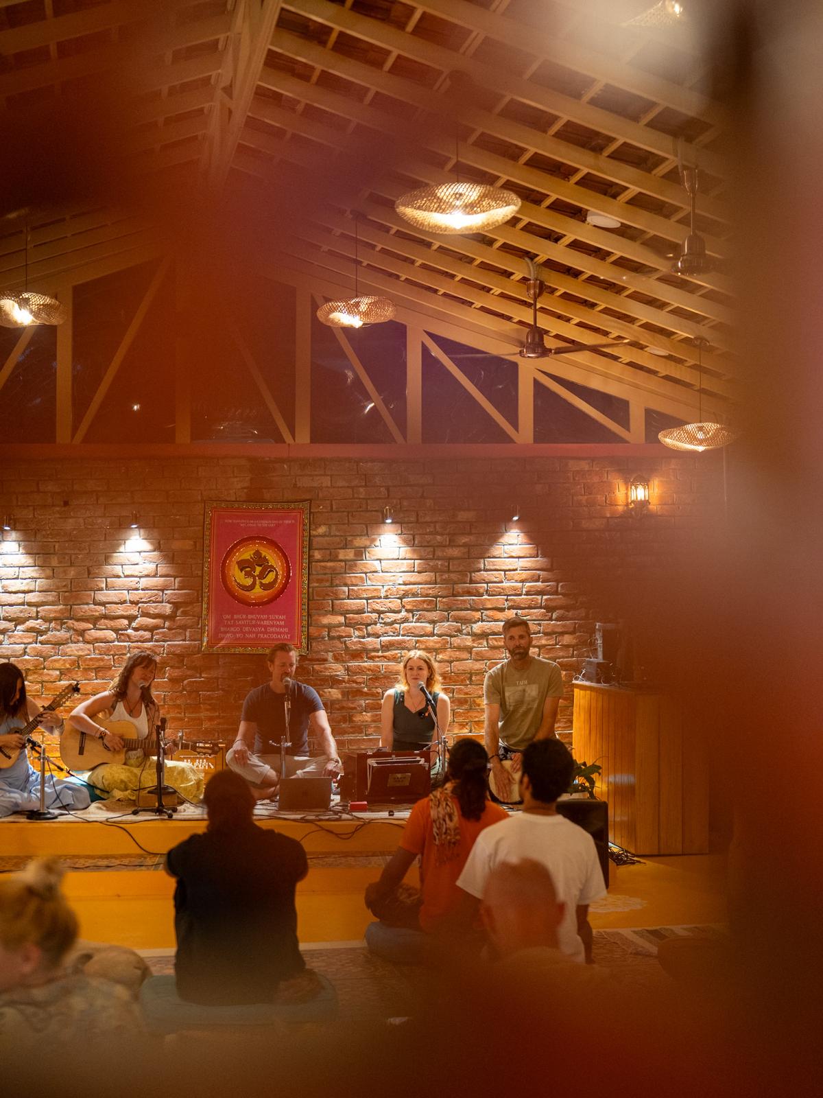 An evening kirtan in the hall, musicians on the low stage under warm lights